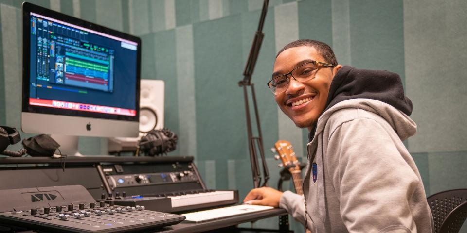 A teen in glasses and a hoodie smiles at the camera while sitting at a recording studio setup with a guitar visible in the background. 