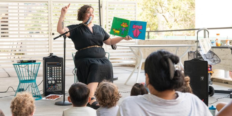 Librarian in a black dress reads a colorful picture book to a group of children and caregivers in an outdoor setting. 
