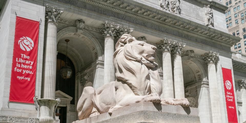 Lion statue in front of the Stephen A. Schwarzman Building with two red banners posted on the facade. 