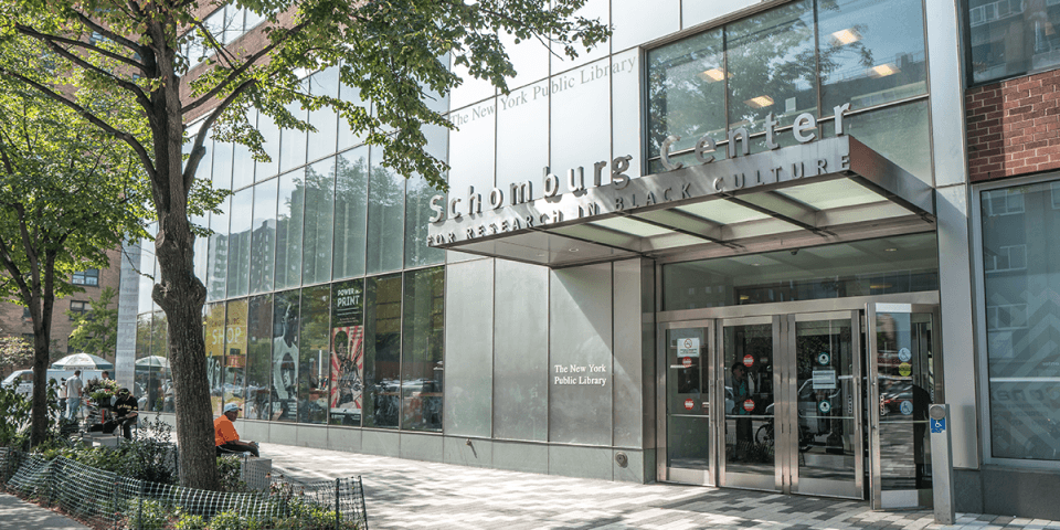 The entrance to the Schomburg Center on a tree-lined sunny street. The Schomburg Center is a multi-story brick building with large windows and chrome paneling.