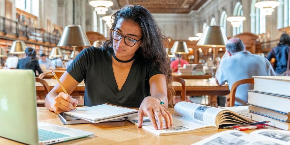 A researcher sits at a table in the Rose Main Reading Room with an open laptop, a notebook, and a stack of books. 