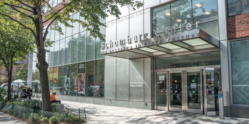 Exterior of the Schomburg Center, featuring large glass windows and a row of lush green trees. 